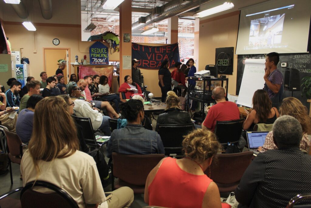 an image showing one person presenting in the front of the room, with about 20 people sitting around listening in a semi-circle. The people in the group are diverse. E4E workshop in Amherst, MA in 2019